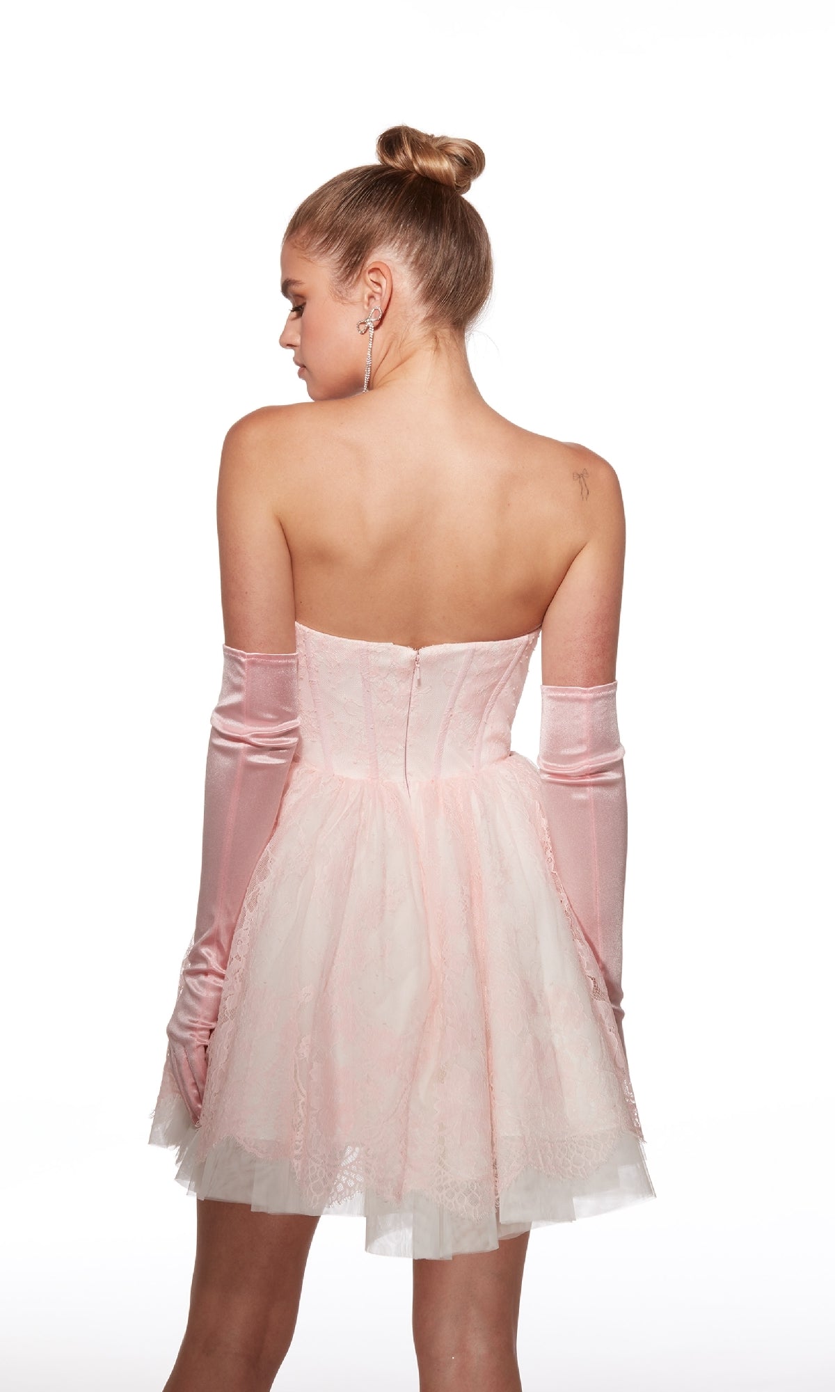 A woman with her hair in a bun wears the strapless short lace hoco dress Alyce 30041 in light pink with matching long gloves, facing away from the camera against a white background.